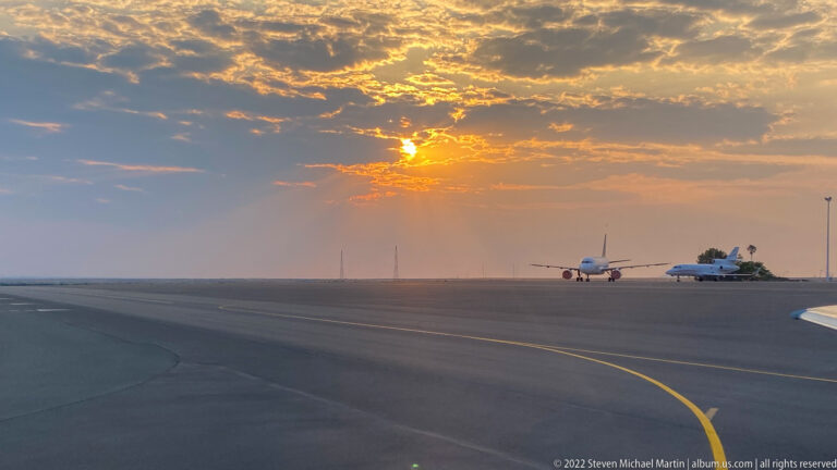 Passenger aircraft stands on a broad airport apron beneath orange sunset clouds, with a curved yellow taxiway line in foreground.