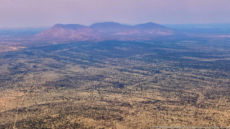 Aerial view of low mountain ridges rising from a broad semi-arid plain patterned with sparse vegetation and drainage lines.