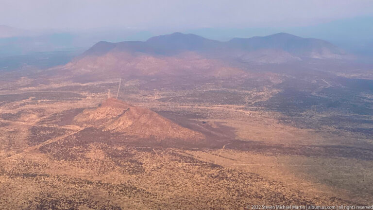 Aerial view of a conical foreground hill and a hazy chain of rounded mountains rising from a broad semi-arid basin.