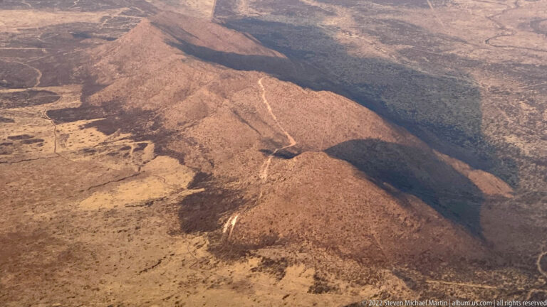 Aerial close view of an eroded rocky ridge with pale runoff scars, dry slopes, and deep shadow along one side.