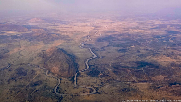 Aerial view of a winding river channel bordered by darker vegetation as it passes between dry hills and plains.