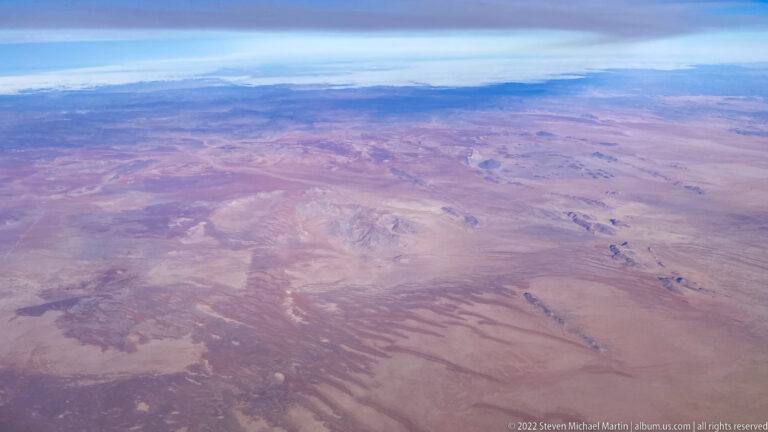 High aerial view of a vast pale desert plain with scattered low ridges, subtle washes, and cloud along the horizon.