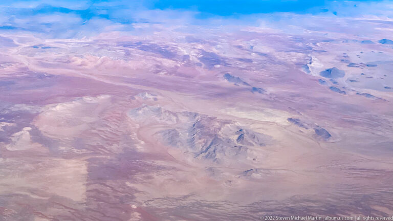 High aerial view of isolated rocky mountain masses rising from a pale pink-beige desert plain beneath patches of blue sky.