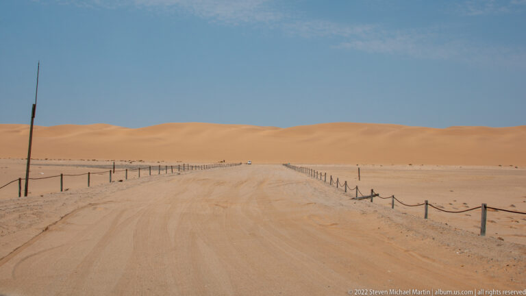 Straight sandy track bordered by posts and rope leading across a pale desert plain toward low dunes.