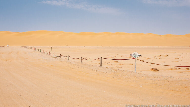 Sandy desert route beside a small diamond sign and rope barriers, with a long orange dune ridge beyond.