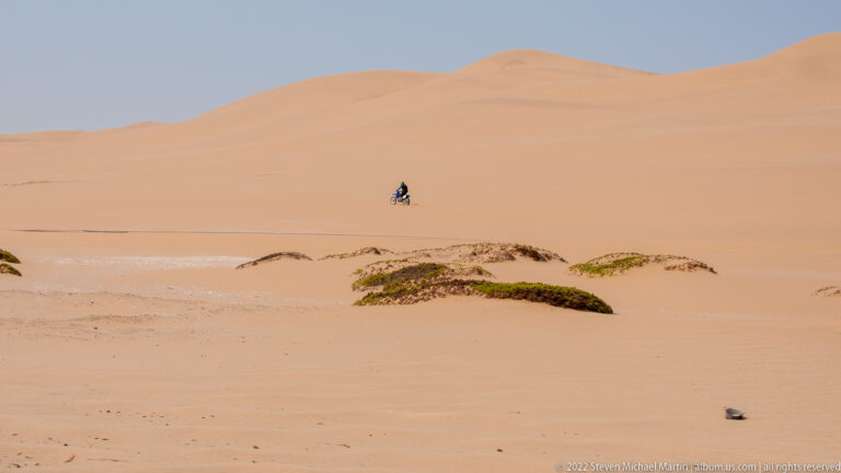 Small lone person crossing a vast pale-orange sand dune, with sparse green vegetation in the foreground.
