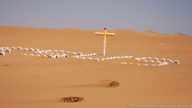 Cross-shaped desert marker encircled by rows of half-buried white tires against orange dunes.