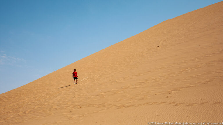 Person in a red shirt descending a steep golden sand dune beneath a clear blue sky.