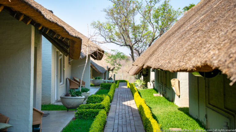 Brick walkway between white lodge buildings with thatched roofs, clipped hedges, shaded verandas, and garden trees.