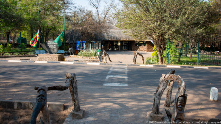 Entrance forecourt with rustic wooden seating, flags, a stone welcome marker, thatched buildings, and several visitors.