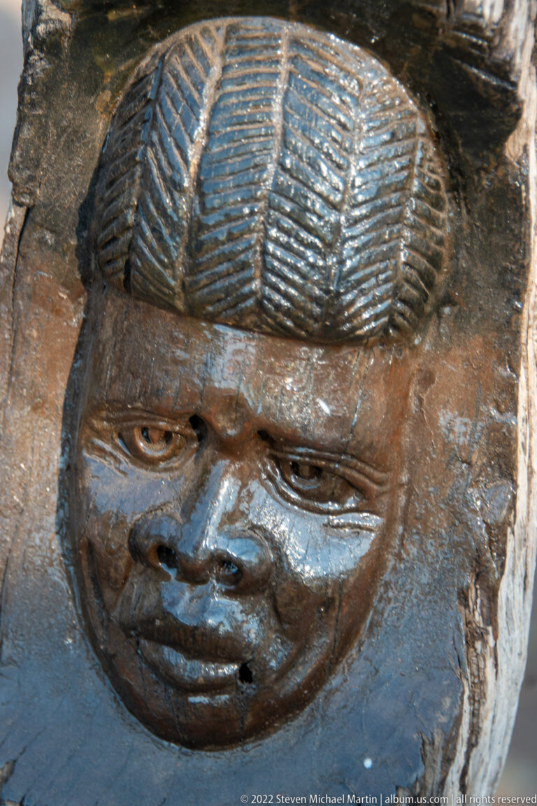 Close-up of a dark wooden carved face with rounded cheeks and a ridged, patterned hairstyle.