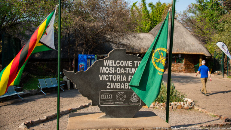 Zimbabwe-shaped welcome marker reading Mosi-oa-Tunya Victoria Falls Zimbabwe, flanked by flags outside a thatched entrance building.