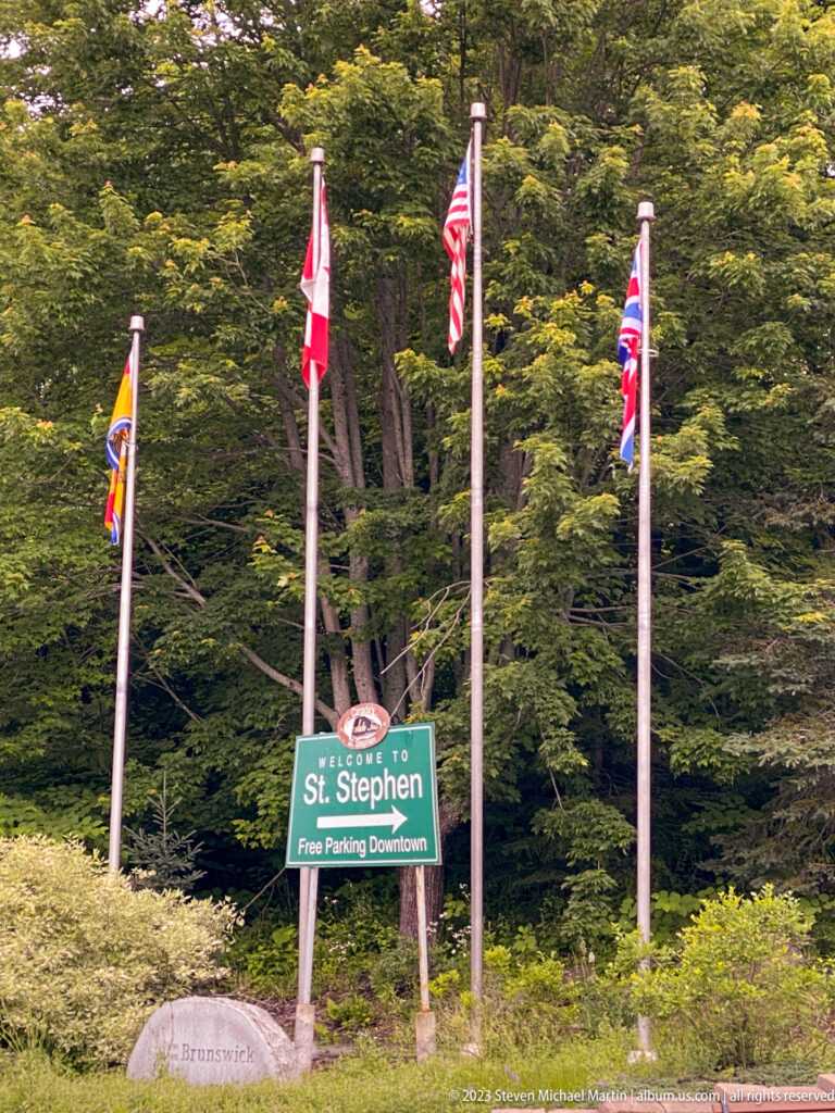 Welcome to St. Stephen sign beneath several flagpoles beside a wooded roadside.