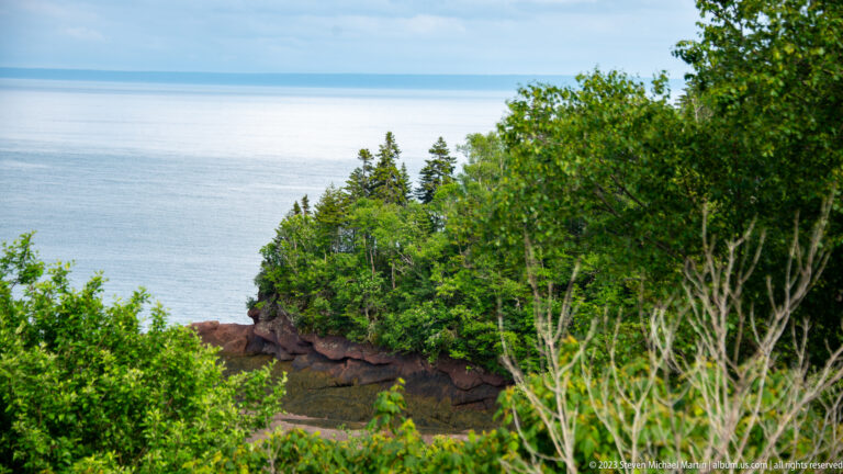 Wooded Herring Cove shoreline with reddish rock ledges and open Bay of Fundy beyond.