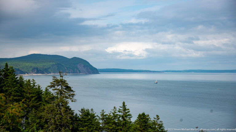 Broad bay view toward a high forested headland under layered clouds, with a small vessel offshore.