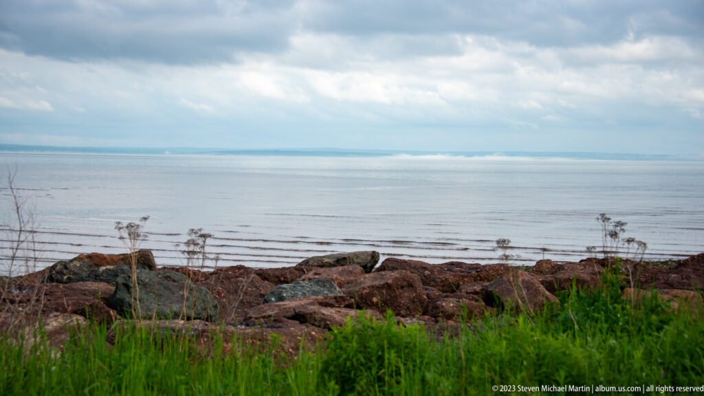 Reddish shoreline rocks and green grasses facing calm Bay of Fundy water beneath a cloudy sky.