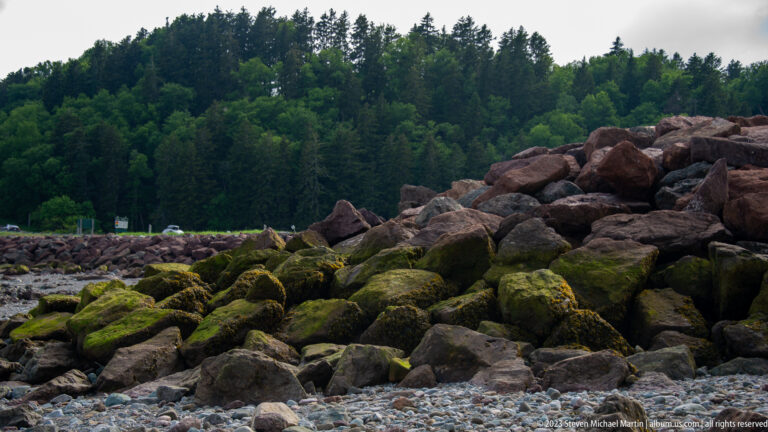 Moss-covered riprap boulders rising from a cobble shore below dense forest.