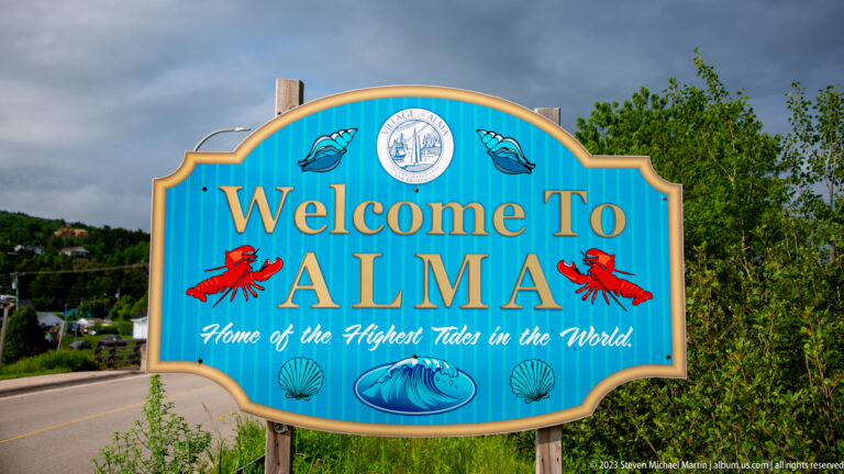 Turquoise Welcome to Alma sign decorated with lobsters, shells, whale, and waves beside the road.