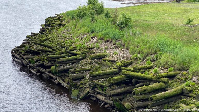 Moss-covered stacked timbers forming old shoreline cribwork along a narrow tidal waterway.