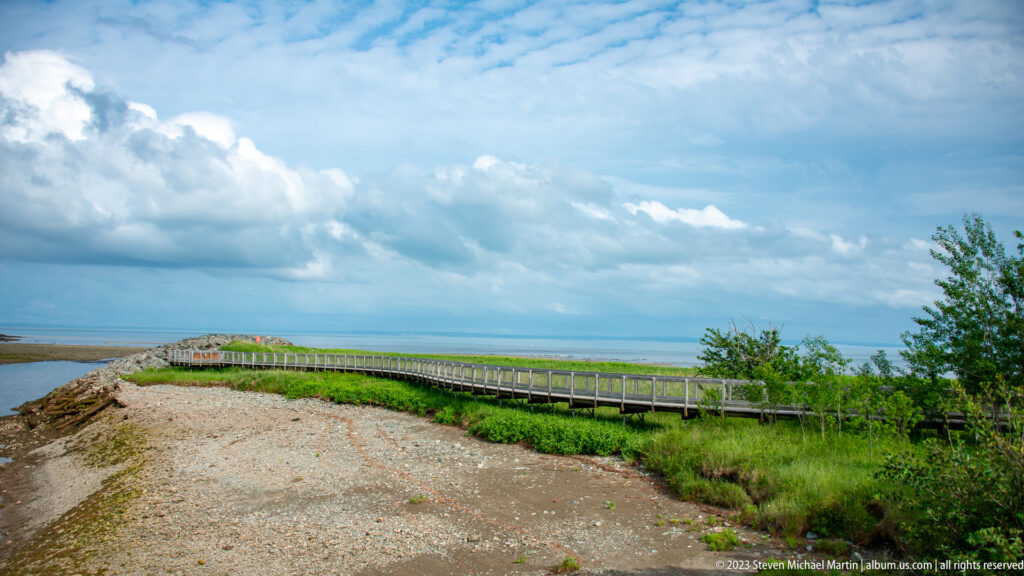 Wooden boardwalk crossing an open coastal overlook above the Bay of Fundy beneath large clouds.