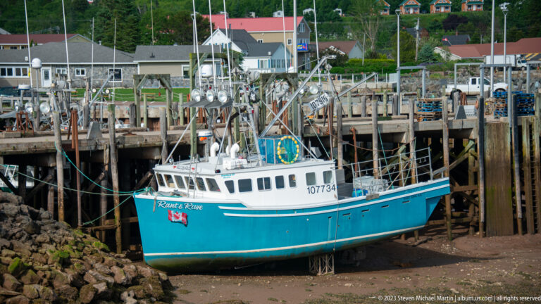 Turquoise fishing boat resting on supports below tall timber wharf pilings at low tide in Alma.