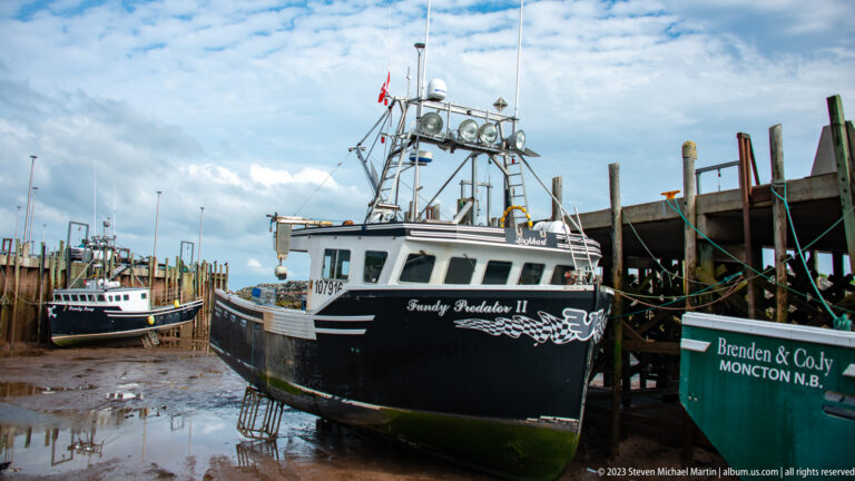 Black fishing vessel Fundy Tradition II resting on supports beside the wharf at low tide.