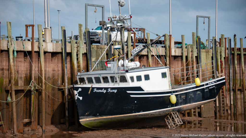 Black-and-white fishing boat Fundy Fury resting on supports below timber wharf pilings at low tide.