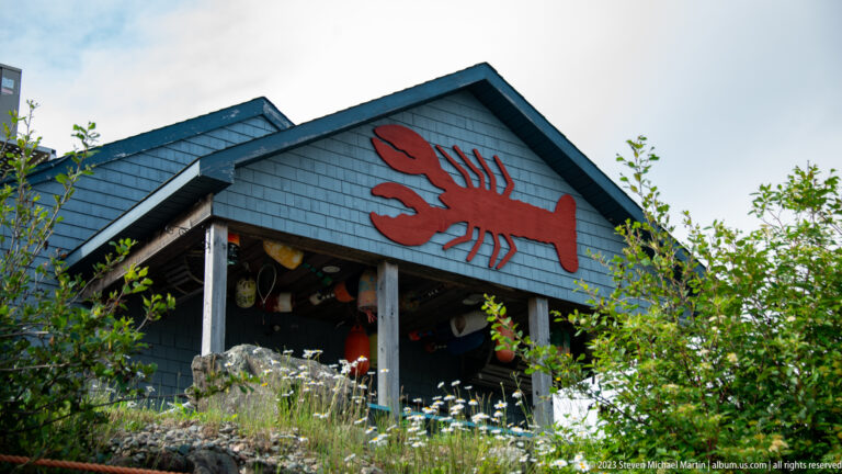 Dark shingled waterfront building with a large red lobster emblem on its gable and maritime objects under the porch.