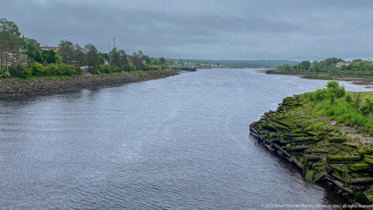 Wide tidal channel bordered by moss-covered timber remains, green banks, and distant houses under gray sky.