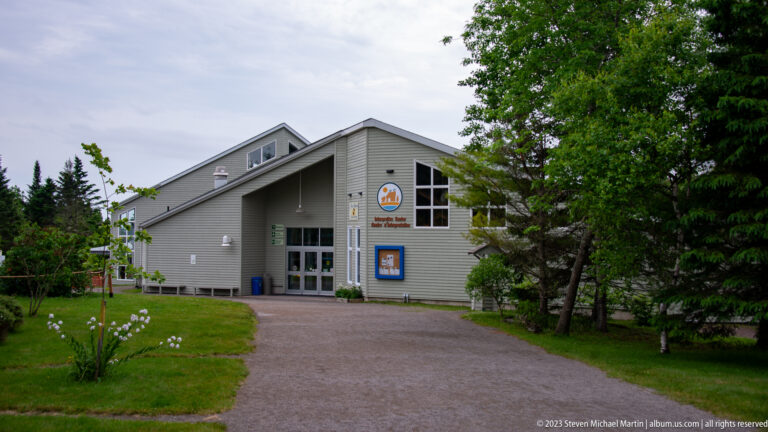 Gray gabled Hopewell Rocks Interpretive Centre approached by a paved path through trees and lawn.