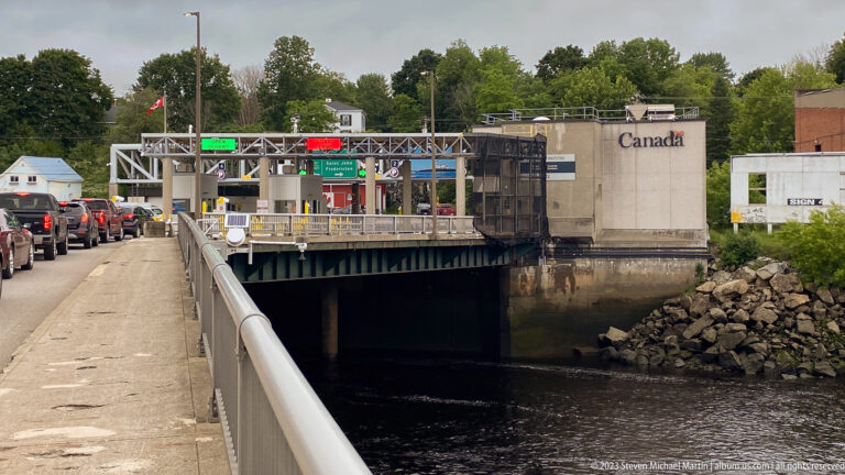Vehicles queue beside a Canada border inspection building and overhead lane signals at a bridge crossing.
