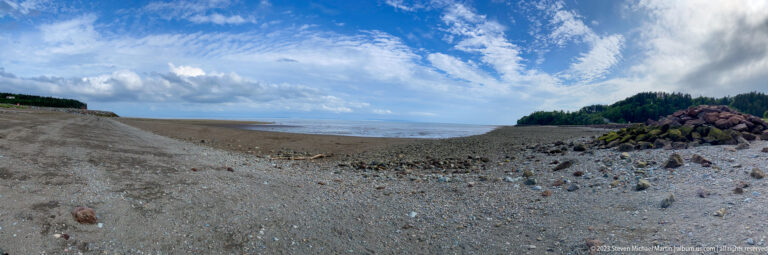 Wide exposed gravel and mud flats near Hopewell Rocks beneath a blue, cloud-filled sky.