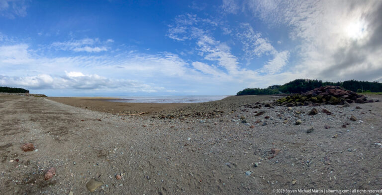 Broad gravelly low-tide shore near Hopewell Rocks with distant water, rocky embankment, and wooded point.