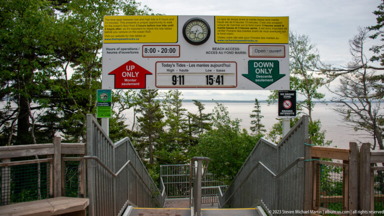 Hopewell Rocks access stairs beneath signs showing hours, directional rules, and the day's high and low tides.