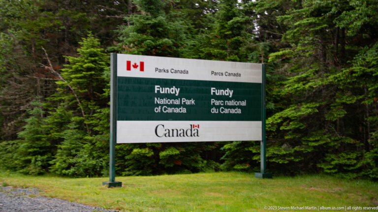 Bilingual Parks Canada sign for Fundy National Park standing on grass in front of evergreen forest.