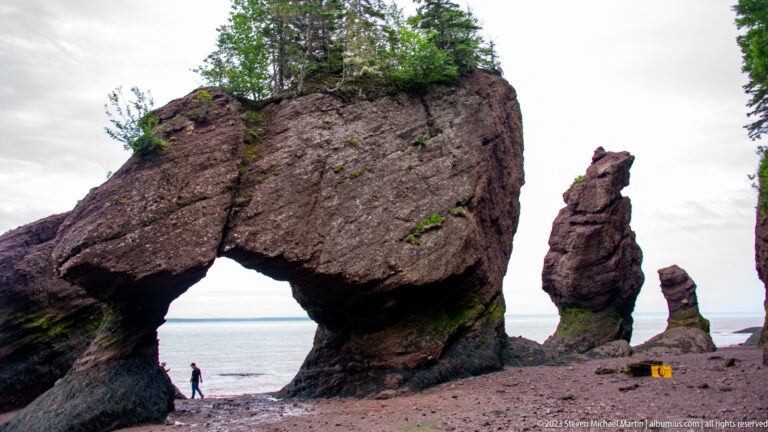 Large Hopewell Rocks natural arch framing the sea, with a visitor beneath it and narrow sea stacks nearby.