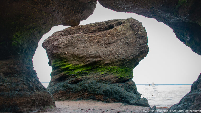 Hopewell flowerpot rock framed through a dark eroded opening in the coastal cliff at low tide.