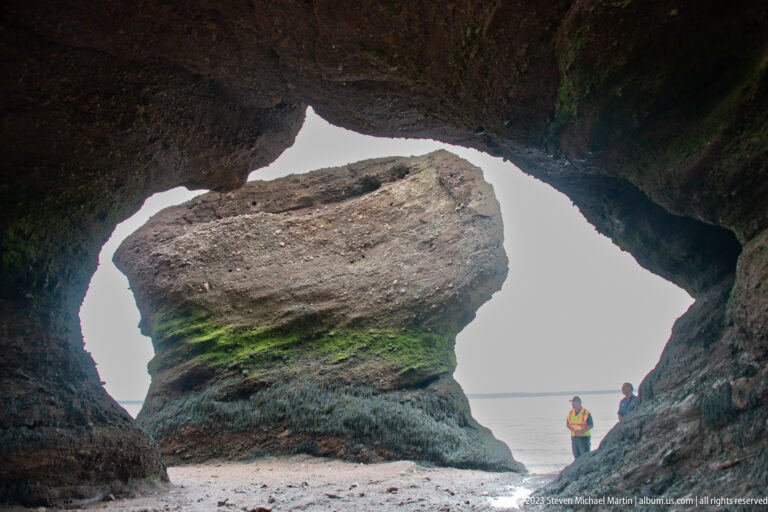 Large Hopewell flowerpot rock framed by a dark opening, with two visitors standing beside it at low tide.