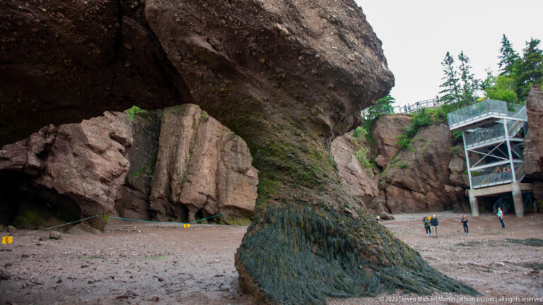 Large dark rock overhang framing the Hopewell Rocks access tower and visitors on the tidal floor.