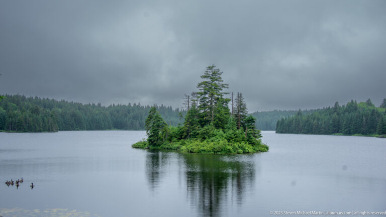 Small evergreen-covered island reflected in a calm lake with forested shores and overcast sky.