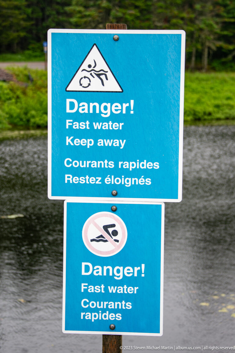 Blue bilingual Danger Fast water signs beside a waterway, including a no-swimming symbol.