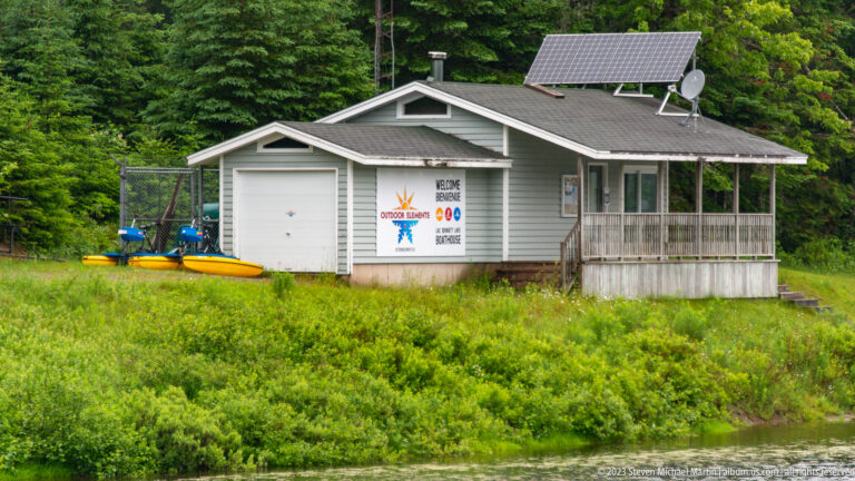 Small gray waterside building with roof solar panels, porch, wall graphics, and stored watercraft beside it.