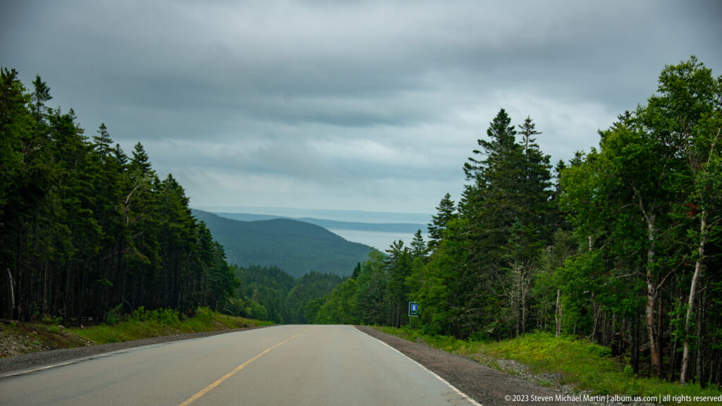 Two-lane road descending between dense forest toward layered distant hills under dark clouds.