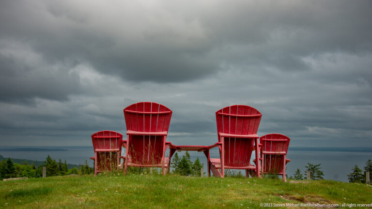 Four red Adirondack chairs on grass facing a distant blue-gray coastal horizon under storm clouds.