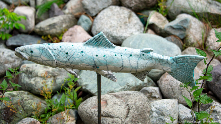 Gray-blue spotted salmon sculpture mounted on a metal support above rounded rocks and low plants.