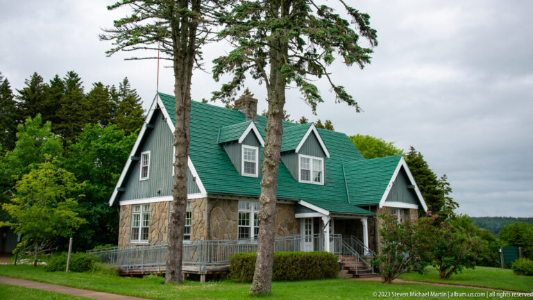 Stone-and-dark-shingle building with steep green roof, white-trimmed dormers, porch, and mature trees.