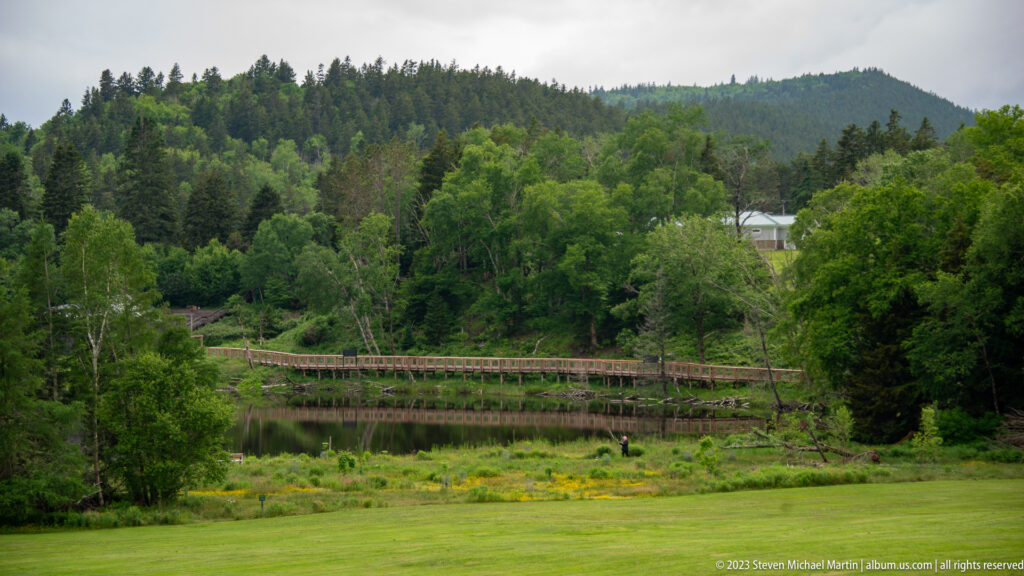 Long wooden walkway crossing a low wet meadow below a forested hillside, viewed across a broad green lawn.