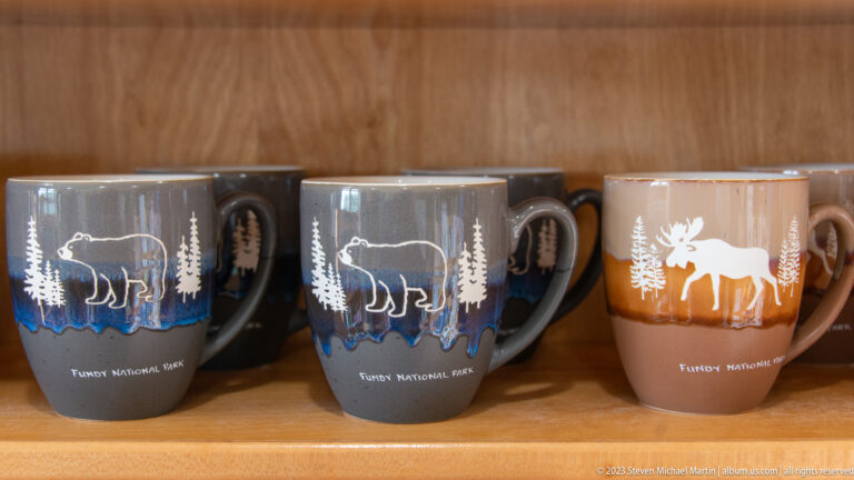 Rows of Fundy National Park souvenir mugs decorated with moose silhouettes and evergreen trees on a wooden shelf.