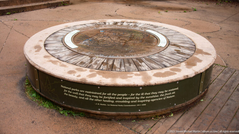 Low circular interpretive table with a central relief map and inscription set into a paved visitor area.