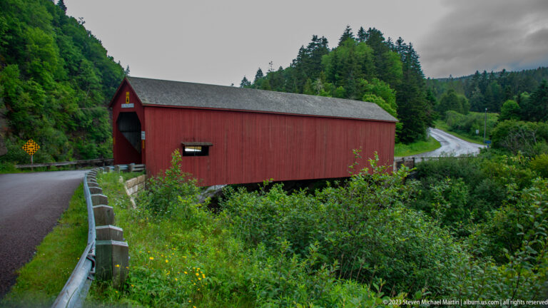 Red Point Wolfe covered bridge spanning a forested rocky gorge beside a curving wet road.